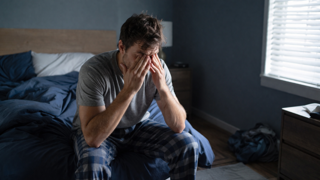 what is sleep debt — tired man sitting on edge of bed exhausted
